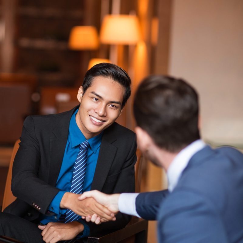 Closeup of two smiling business men shaking hands in cafe. One man is sitting back to camera.